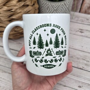 White ceramic mug with green forest‑school design featuring pine trees, campfire, logs, boots, and the phrase “Not all classrooms have four walls – Forest School,” held in a hand above a light wooden surface beside a woven basket and grey patterned plant pot.