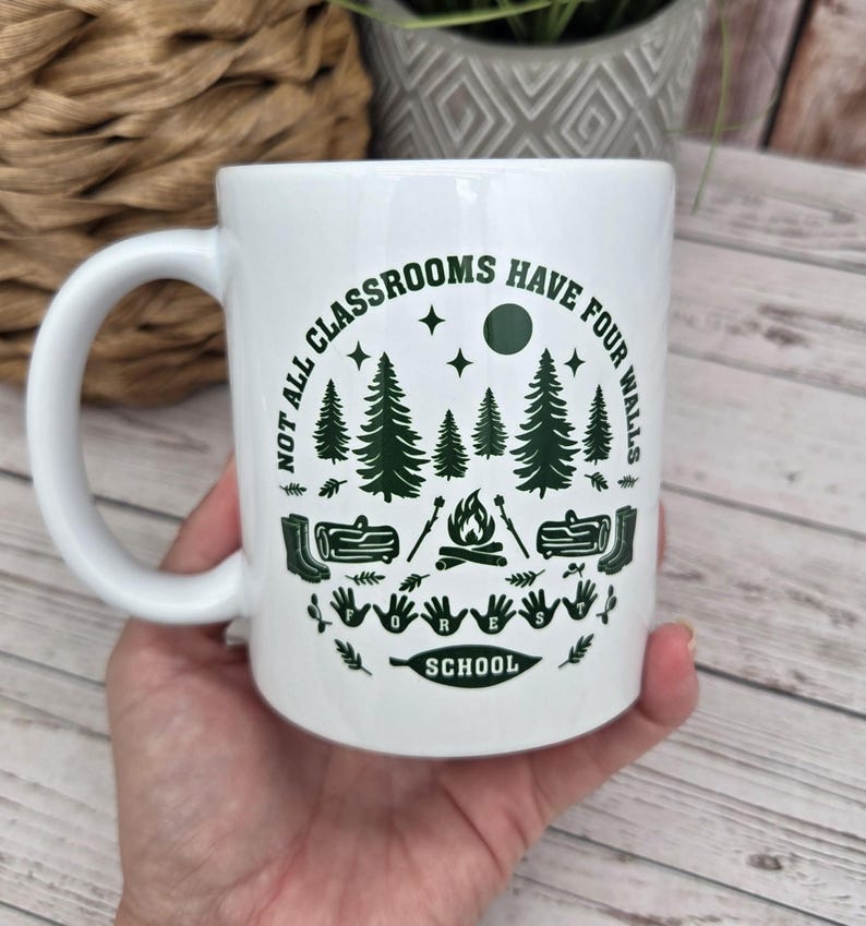 White ceramic mug with green forest‑school design featuring pine trees, campfire, logs, boots, and the phrase “Not all classrooms have four walls – Forest School,” held in a hand above a light wooden surface beside a woven basket and grey patterned plant pot.