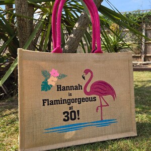 Natural colour jute bag with pink handle. Displays a pink flamingo using glitter htv, standing in a glitter blue lake, with flowers to one side, personalised with Hannah is Flamingorgeous at 30!