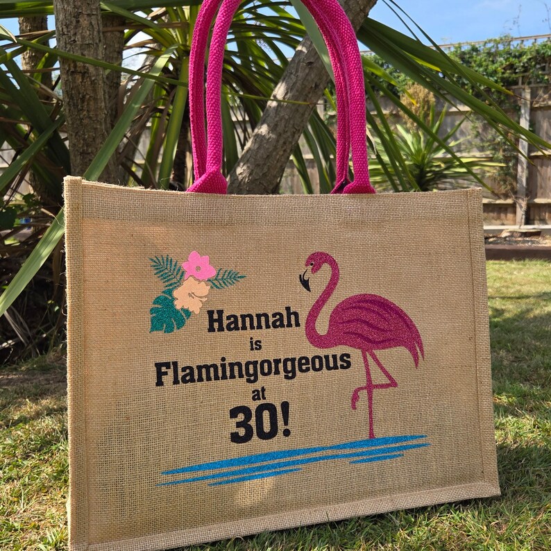 Natural colour jute bag with pink handle. Displays a pink flamingo using glitter htv, standing in a glitter blue lake, with flowers to one side, personalised with Hannah is Flamingorgeous at 30!