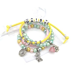 Può includere: Un set di braccialetti di perline dai colori pastello, tra cui rosa, giallo e verde. I braccialetti hanno ciondoli, tra cui un coniglietto, un fiore e un uovo di Pasqua. La parola "DAISY" è scritta con perline bianche su un cordino giallo.