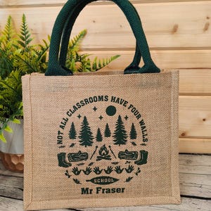 A Jute tote bag with green handles. Each bag features a circular design with trees, a campfire, and the text "NOT ALL CLASSROOMS HAVE FOUR WALLS SCHOOL". With the name Mr Fraser