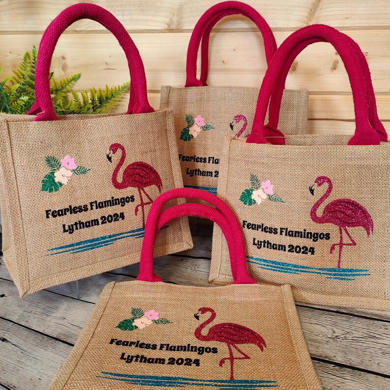 Four small burlap tote bags with pink flamingo designs and the text "Fearless Flamingos Lytham 2024". The bags have red handles.
