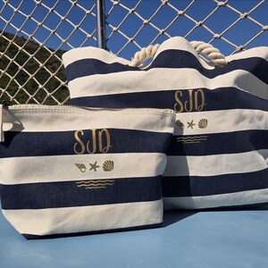 Puede incluir: Un bolso tote a rayas azul marino y blanco y una bolsa con cremallera a juego. Ambos artículos presentan iniciales doradas, una estrella de mar, una concha y un diseño de olas. El bolso tote tiene asas de cuerda.