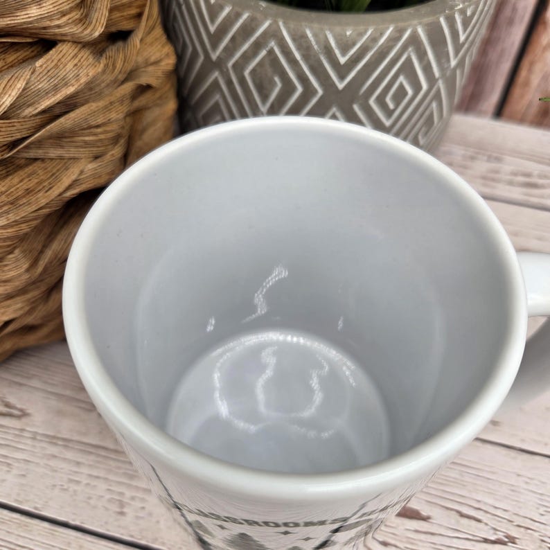 Close‑up of the inside of a white ceramic mug showing its glossy, clean interior, placed on a light wooden surface next to a grey geometric plant pot and woven basket.