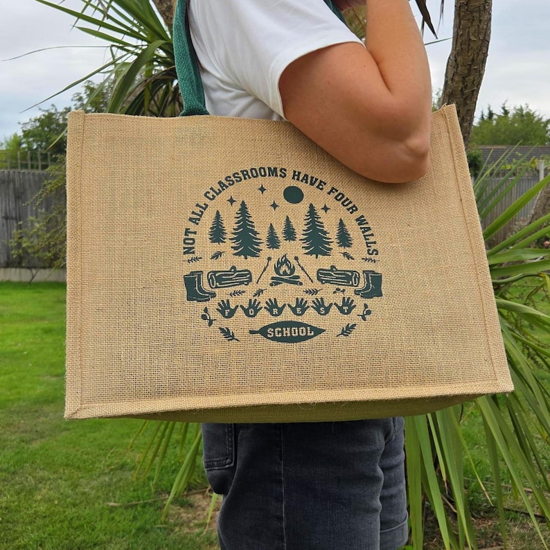 Person holding a large jute tote bag with teal rope handles outdoors. The bag displays a dark green design of trees, logs, a campfire, and handprints with the text “Not all classrooms have four walls – School.” The background includes green grass, plants, and a wooden fence.