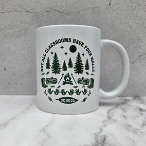 White ceramic mug with dark green forest‑school design featuring pine trees, campfire, logs, boots, and the phrase “Not all classrooms have four walls – Forest School,” placed on a marble surface against a grey concrete background.