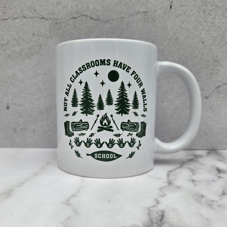White ceramic mug with dark green forest‑school design featuring pine trees, campfire, logs, boots, and the phrase “Not all classrooms have four walls – Forest School,” placed on a marble surface against a grey concrete background.