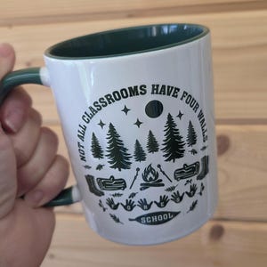 White mug with dark green interior and handle, featuring forest‑school design with trees, campfire, and boots, held against a wooden wall background.