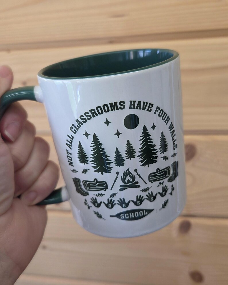 White mug with dark green interior and handle, featuring forest‑school design with trees, campfire, and boots, held against a wooden wall background.