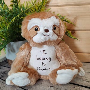 May include: A soft toy sloth with brown and white fur, embroidered with the words "I belong to Nancy". The sloth has a smiling face and white paws. The toy is positioned in front of a green plant.
