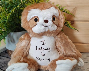 May include: A plush sloth toy with brown and white fur, embroidered with the words "I belong to Nancy". The sloth has a smiling face and white paws. The toy is sitting in front of a green plant.