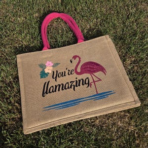 Natural colour jute bag with pink handle. Displays a pink flamingo using glitter htv, standing in a glitter blue lake, with flowers to one side, personalised with your flamazing