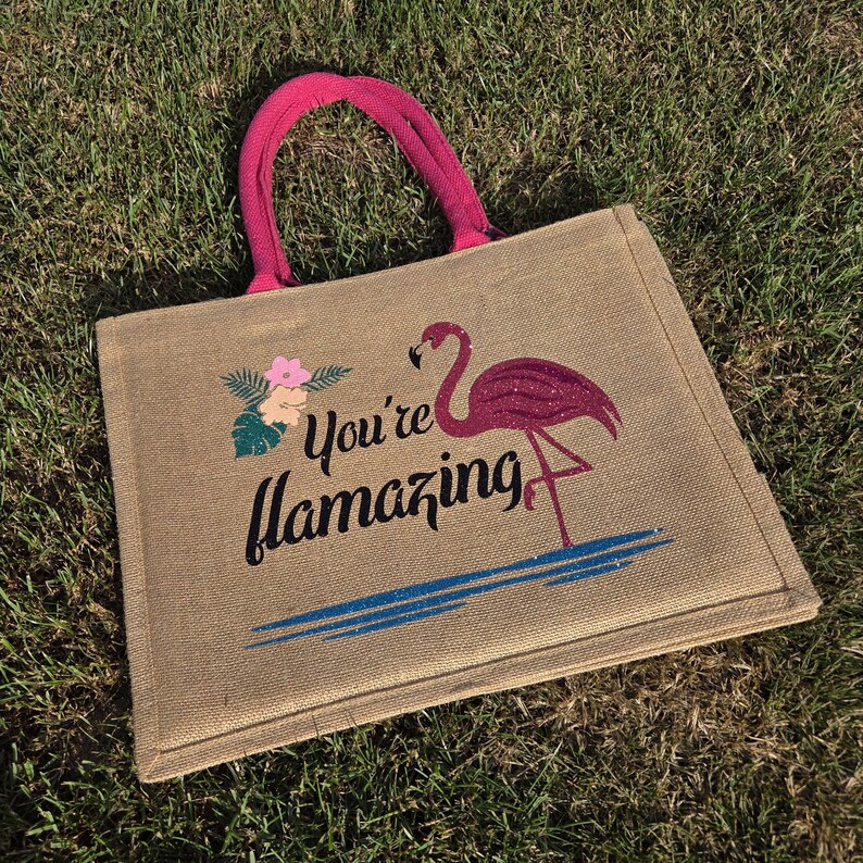 Natural colour jute bag with pink handle. Displays a pink flamingo using glitter htv, standing in a glitter blue lake, with flowers to one side, personalised with your flamazing