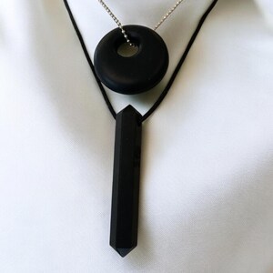 Puede incluir: Un collar negro con dos colgantes. Un colgante es un c&iacute;rculo redondo y plano con un agujero en el centro. El otro colgante es una forma larga, delgada y puntiaguda.