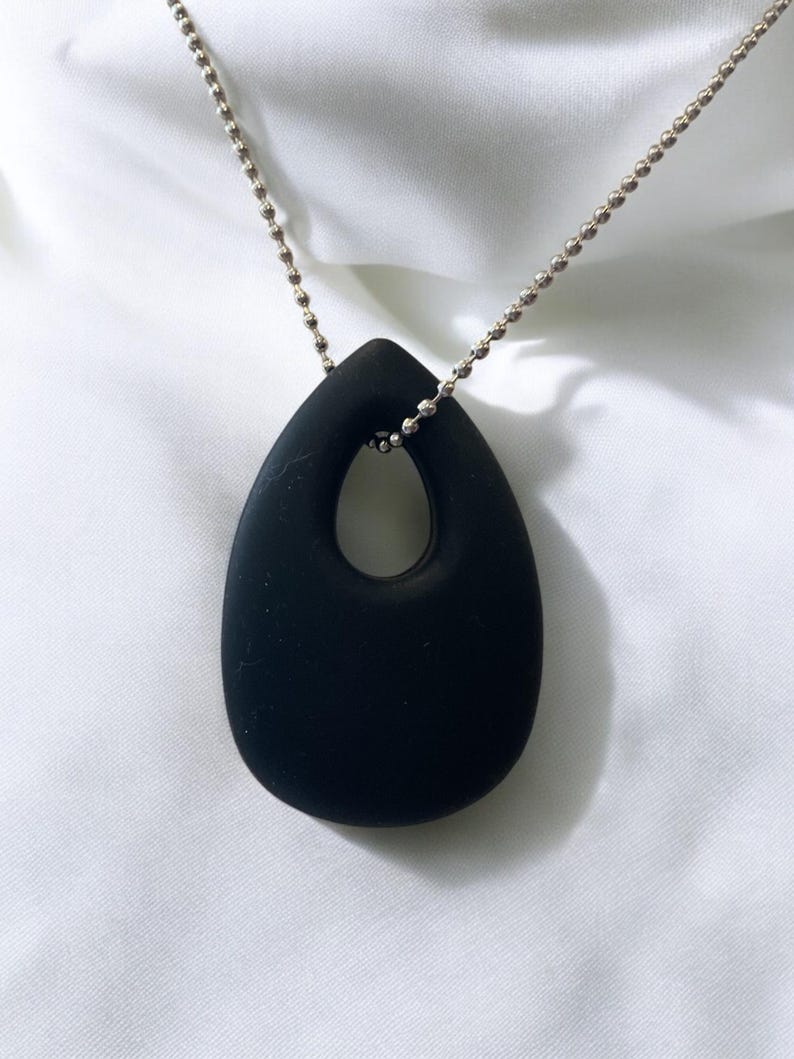 Puede incluir: Un collar con un colgante negro en forma de l&aacute;grima con un gran recorte ovalado. El colgante est&aacute; unido a una cadena plateada.