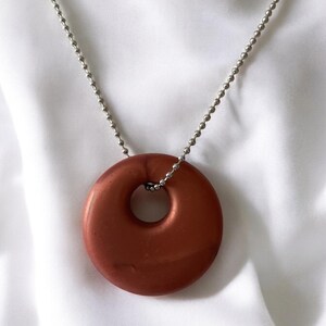 May include: A copper-colored pendant necklace with a circular donut-shaped pendant. The pendant hangs from a silver ball chain. The background is a white, silky fabric.