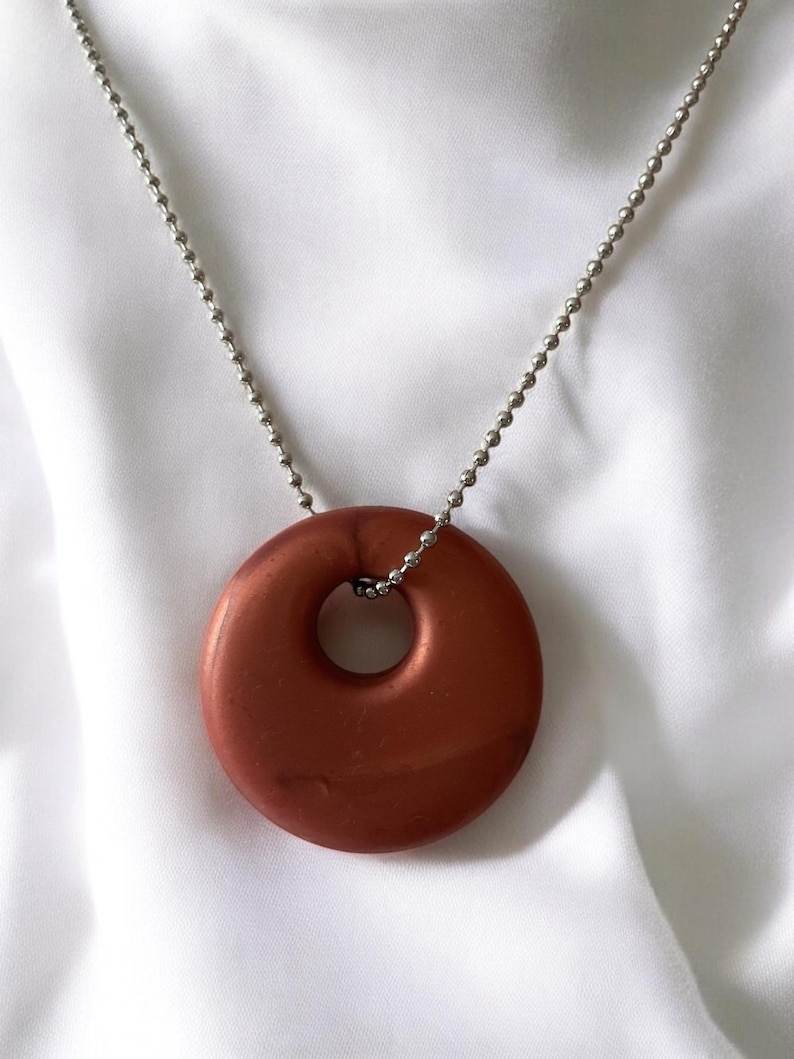 May include: A copper-colored pendant necklace with a circular donut-shaped pendant. The pendant hangs from a silver ball chain. The background is a white, silky fabric.