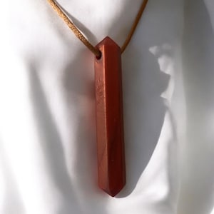 May include: A reddish-brown crystal pendant necklace. The pendant is a long, rectangular shape with a pointed end and a small hole at the top. It hangs from a thin, tan cord. The pendant has a hand symbol etched on it.