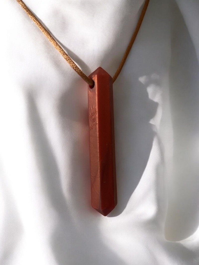 May include: A reddish-brown crystal pendant necklace. The pendant is a long, rectangular shape with a pointed end and a small hole at the top. It hangs from a thin, tan cord. The pendant has a hand symbol etched on it.