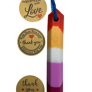 May include: A colorful pendant with a blue cord, featuring a layered design in red, orange, yellow, white, pink, and magenta. The pendant has a geometric shape. Three round stickers with handwritten text and hearts are also visible.