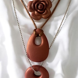 May include: A layered necklace featuring three pendants in a copper-bronze hue. The top pendant is a rose, followed by a teardrop shape, and a circular pendant at the bottom. The necklace has a brown cord and a silver ball chain.