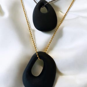 Puede incluir: Dos collares colgantes de cer&aacute;mica negra con una forma ovalada &uacute;nica y un agujero en el centro. El collar superior est&aacute; en una cadena negra y el collar inferior est&aacute; en una cadena dorada.