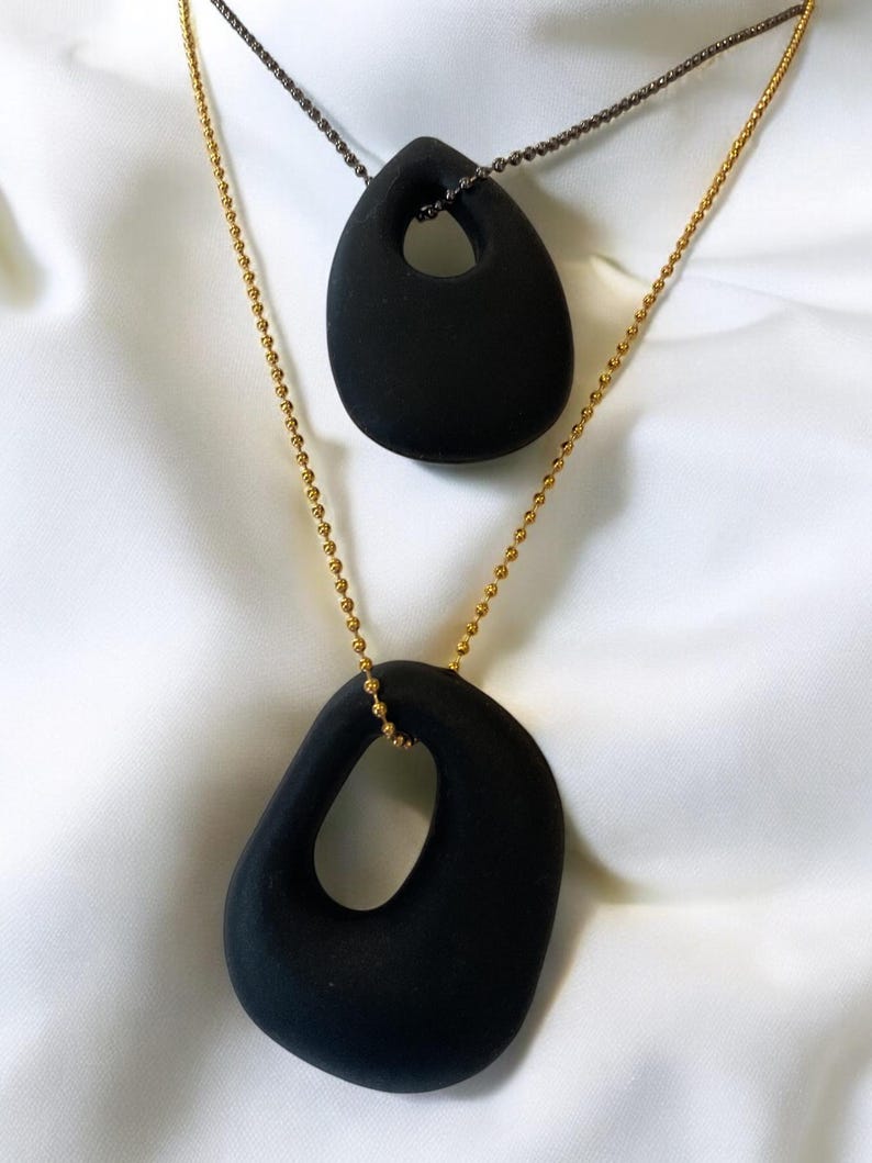 Puede incluir: Dos collares colgantes de cer&aacute;mica negra con una forma ovalada &uacute;nica y un agujero en el centro. El collar superior est&aacute; en una cadena negra y el collar inferior est&aacute; en una cadena dorada.