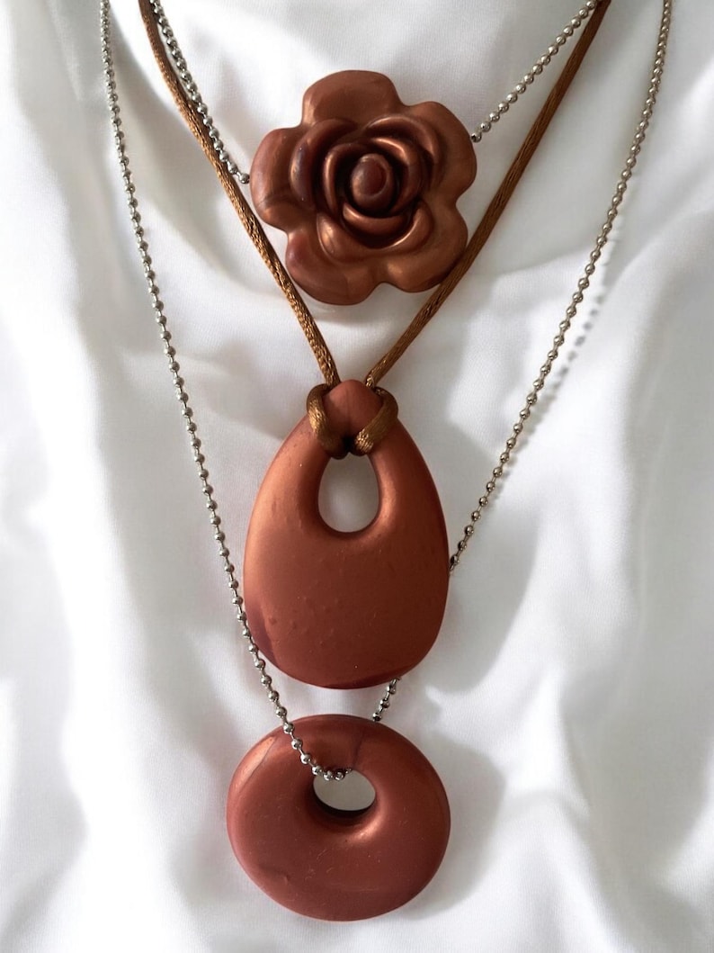 May include: A layered necklace featuring three pendants in a copper-colored finish. The top pendant is a rose, the middle is a teardrop shape, and the bottom is a ring. The necklace has a brown cord and a silver ball chain.
