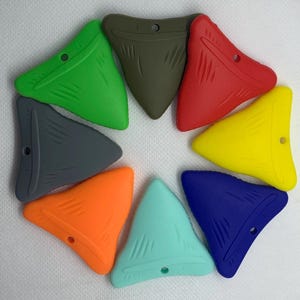 Può includere: Una collezione di otto massaggiagengive in silicone a forma di dente di squalo, disposti a cerchio. I massaggiagengive sono in vari colori, tra cui verde, grigio, arancione, verde acqua, rosso, giallo, blu e verde oliva. Ogni massaggiagengive ha un piccolo foro.