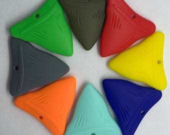 May include: A collection of eight colorful shark tooth-shaped silicone teethers in a circular arrangement. The teethers are in various colors, including green, gray, orange, teal, red, yellow, blue, and olive green. Each teether has a small hole.