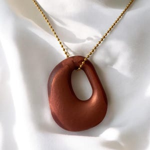 May include: A brown pendant necklace with a gold-colored chain. The pendant is an abstract, organic shape with a smooth, glossy finish. The necklace is displayed on a white, silky fabric.