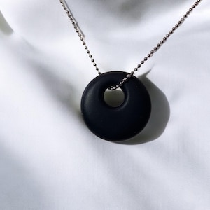 Puede incluir: Un collar con un colgante redondo negro en forma de donut. El colgante tiene un agujero en el centro. El collar est&aacute; hecho de una cadena plateada.