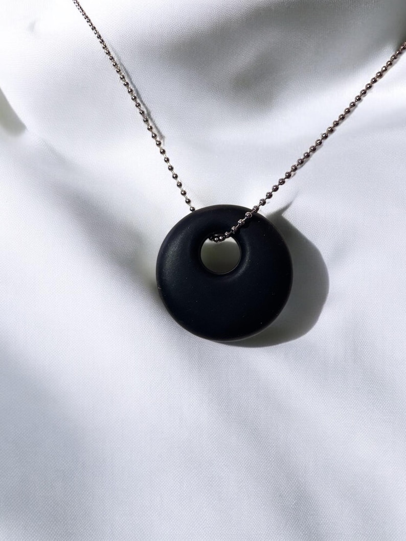 Puede incluir: Un collar con un colgante redondo negro en forma de donut. El colgante tiene un agujero en el centro. El collar est&aacute; hecho de una cadena plateada.