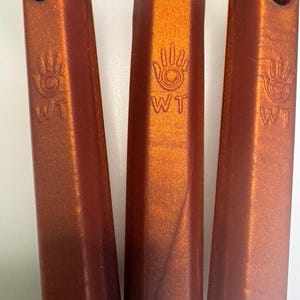 May include: Three metallic orange pendants with a handprint design and the letters "WT" are displayed. Each pendant is rectangular with a faceted surface and a hole at the top for a cord. The cords are brown and gold.