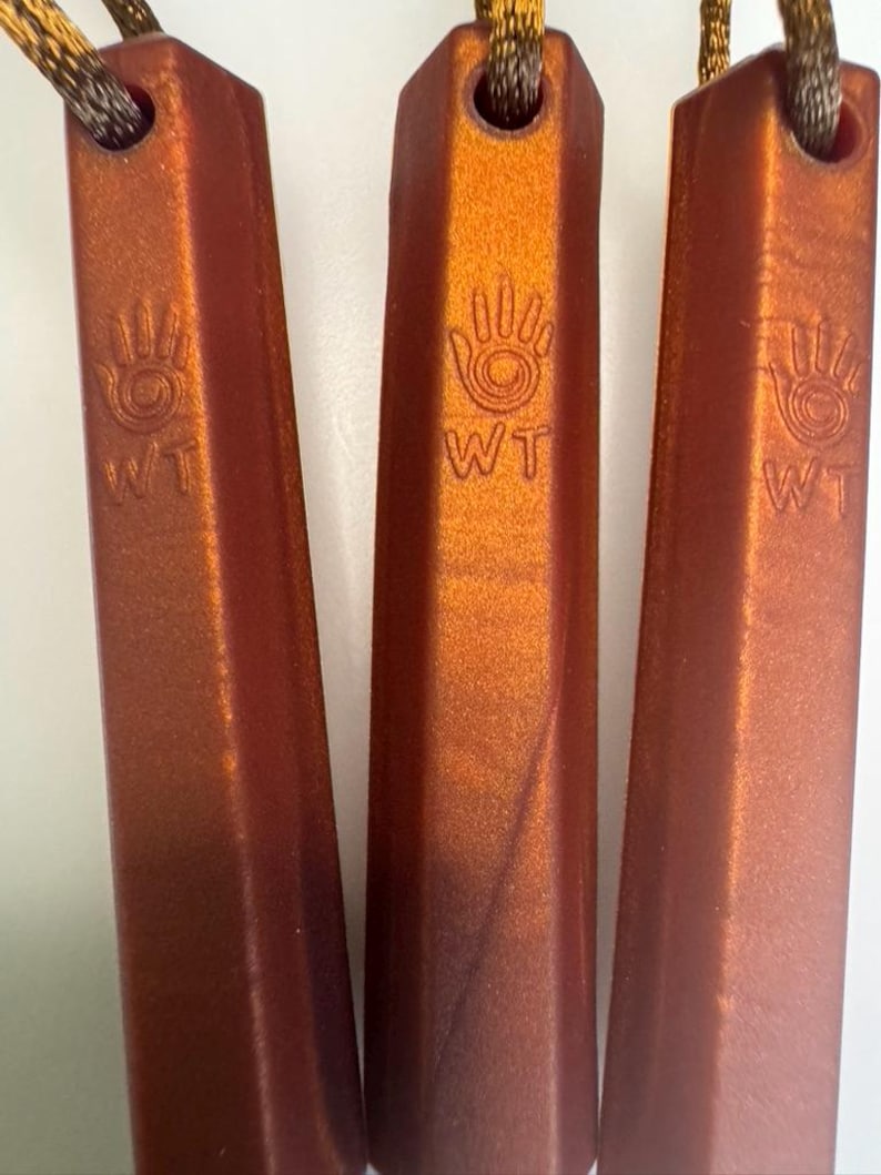 May include: Three metallic orange pendants with a handprint design and the letters "WT" are displayed. Each pendant is rectangular with a faceted surface and a hole at the top for a cord. The cords are brown and gold.