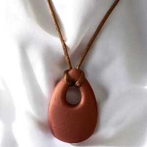 May include: A copper-colored pendant necklace with a teardrop-shaped pendant. The pendant has a large central opening and hangs from a thin, brown cord. The necklace is displayed on a white, textured surface.