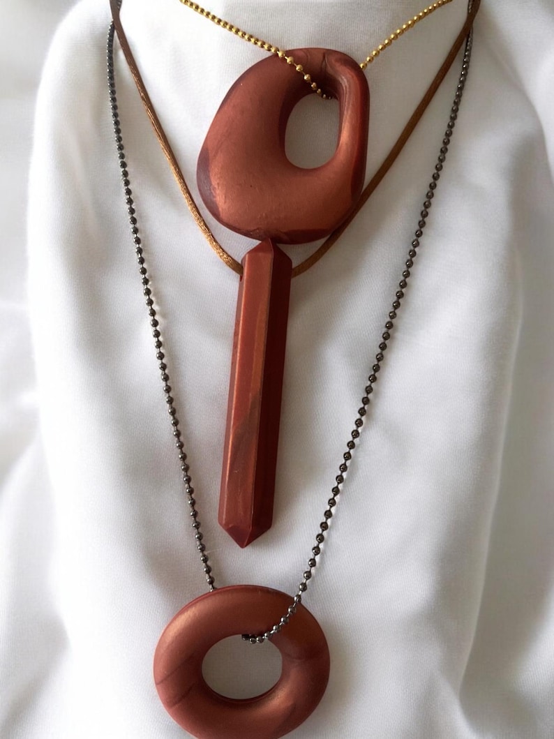 May include: Three necklaces with copper-colored pendants. The top necklace has a large, irregular pendant with a hole. The middle necklace has a long, rectangular pendant. The bottom necklace has a circular pendant.