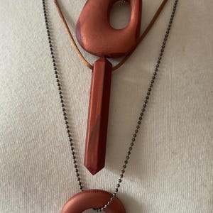 May include: Three necklaces with reddish-brown pendants. The top necklace has a large, irregular pendant with a hole. The middle necklace has a long, rectangular pendant. The bottom necklace has a circular pendant. The chains are gold, brown, and dark gray.