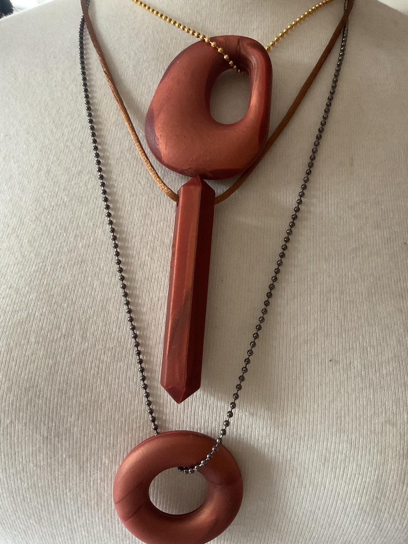 May include: Three necklaces with reddish-brown pendants. The top necklace has a large, irregular pendant with a hole. The middle necklace has a long, rectangular pendant. The bottom necklace has a circular pendant. The chains are gold, brown, and dark gray.