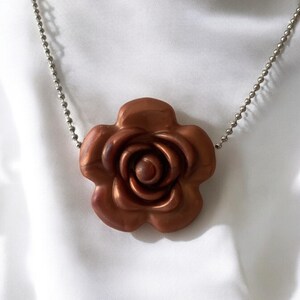 May include: A copper-colored rose pendant necklace. The rose pendant is detailed with multiple petals and a central spiral. The necklace chain is a silver-colored ball chain. The background is a white, silky fabric.