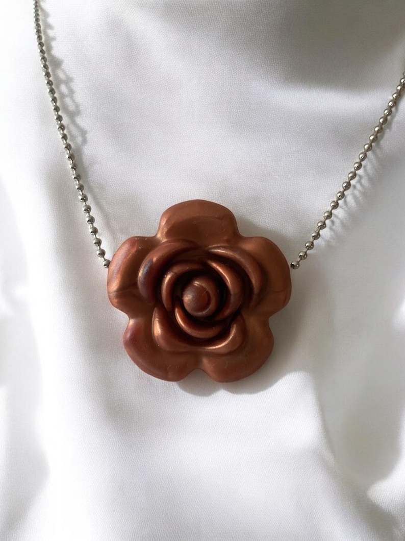 May include: A copper-colored rose pendant necklace. The rose pendant is detailed with multiple petals and a central spiral. The necklace chain is a silver-colored ball chain. The background is a white, silky fabric.