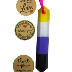 May include: A non-binary pride flag pendant with a yellow, white, purple, and black design, suspended from a pink cord. The pendant has a hexagonal shape. Three brown stickers with text and hearts are also visible.