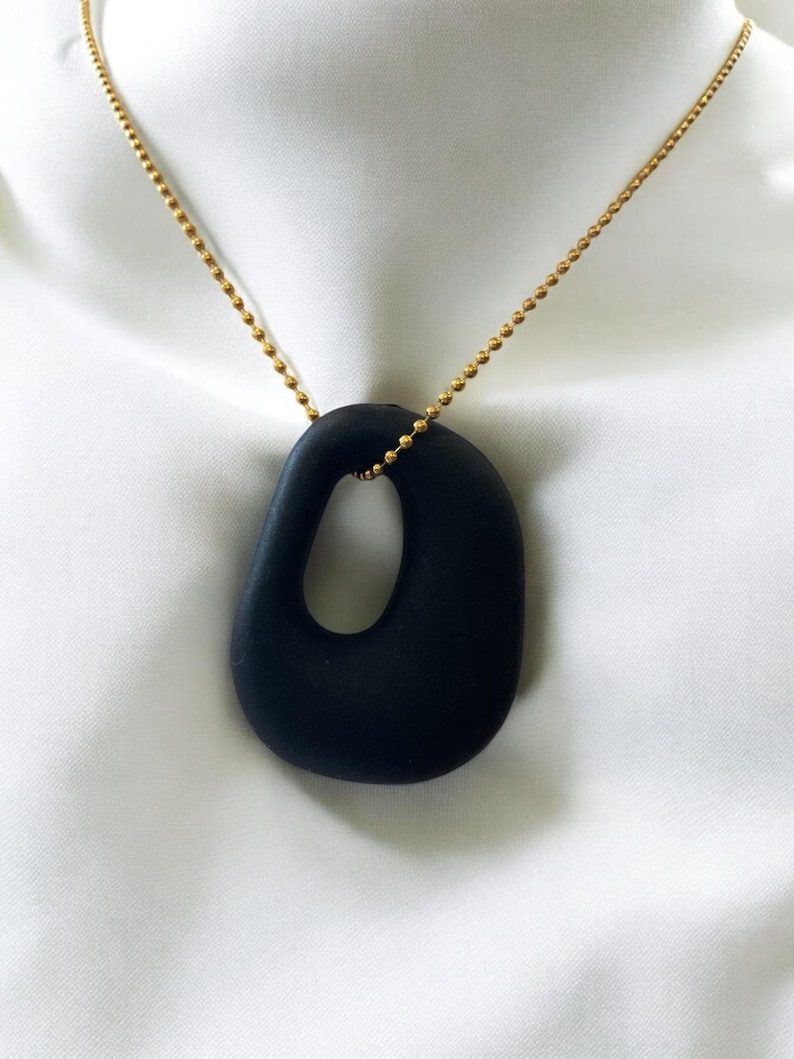 Puede incluir: Un collar de cadena dorada con un colgante ovalado negro y liso, que presenta un c&iacute;rculo grande abierto en el centro.