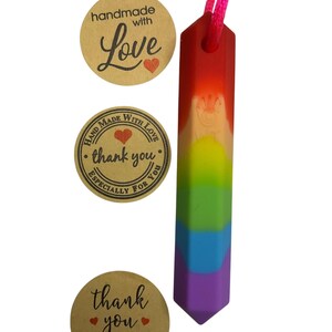 May include: A rainbow-colored sensory chew necklace with a pink cord. The pendant is a rectangular prism with a rounded top and a pointed bottom. Three round stickers with the text "handmade with love" and "thank you" are also visible.