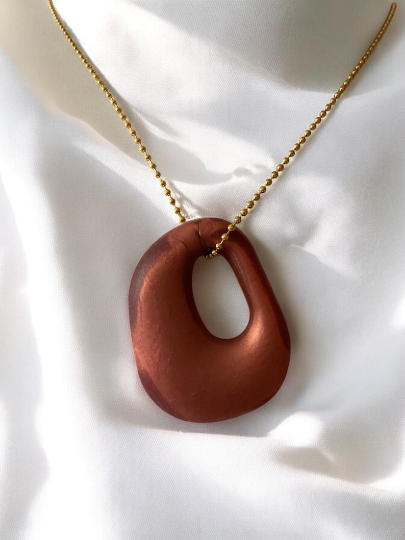 May include: A brown pendant necklace with a gold-colored chain. The pendant is an abstract, organic shape with a smooth, glossy finish. The necklace is displayed on a white, silky fabric.