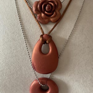 May include: A three-strand necklace set. The top strand features a copper-colored rose pendant on a brown cord. The middle strand has a copper-colored teardrop pendant, and the bottom strand has a copper-colored donut pendant. All pendants are strung on silver ball chains.