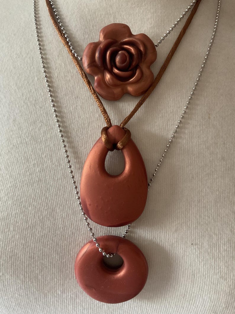 May include: A three-strand necklace set. The top strand features a copper-colored rose pendant on a brown cord. The middle strand has a copper-colored teardrop pendant, and the bottom strand has a copper-colored donut pendant. All pendants are strung on silver ball chains.
