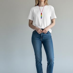 May include: A white t-shirt and blue jeans are worn with a rainbow-colored pendant necklace. The necklace has a red cord and a rectangular pendant with vertical stripes of rainbow colors. The model is wearing white sneakers.
