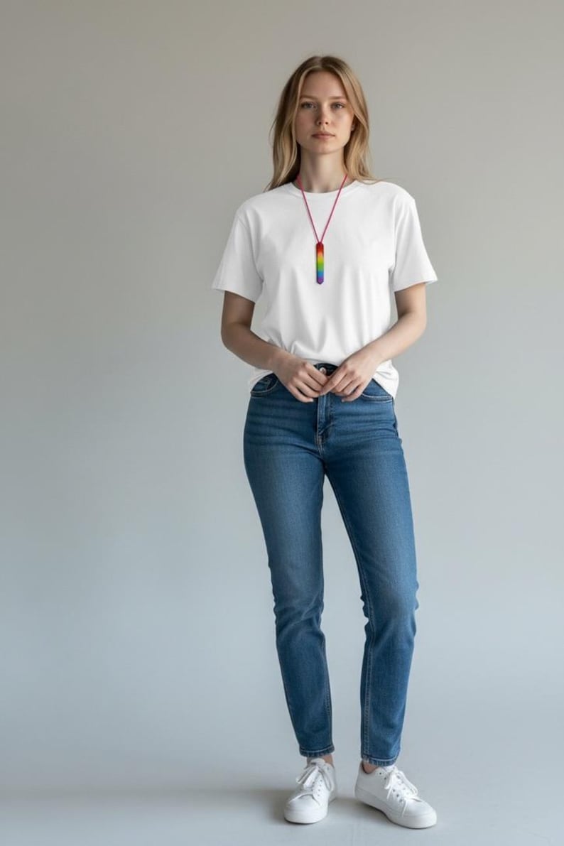 May include: A white t-shirt and blue jeans are worn with a rainbow-colored pendant necklace. The necklace has a red cord and a rectangular pendant with vertical stripes of rainbow colors. The model is wearing white sneakers.